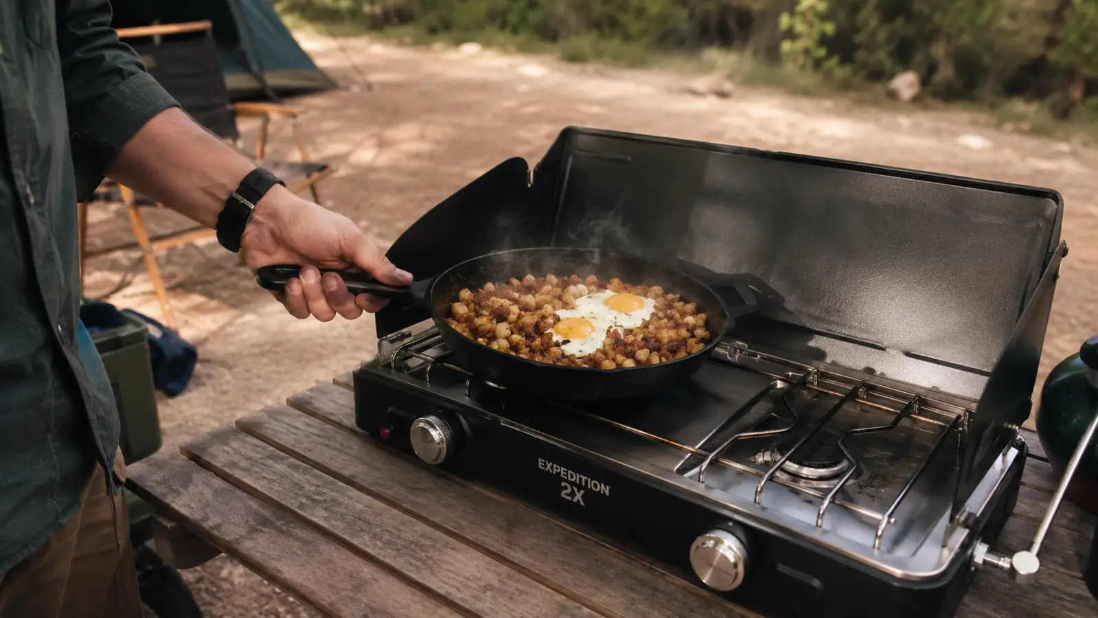 A hand setting the skillet onto a portable two-burner camp stove, hash and eggs visible in the pan, an outdoor table in soft daylight.