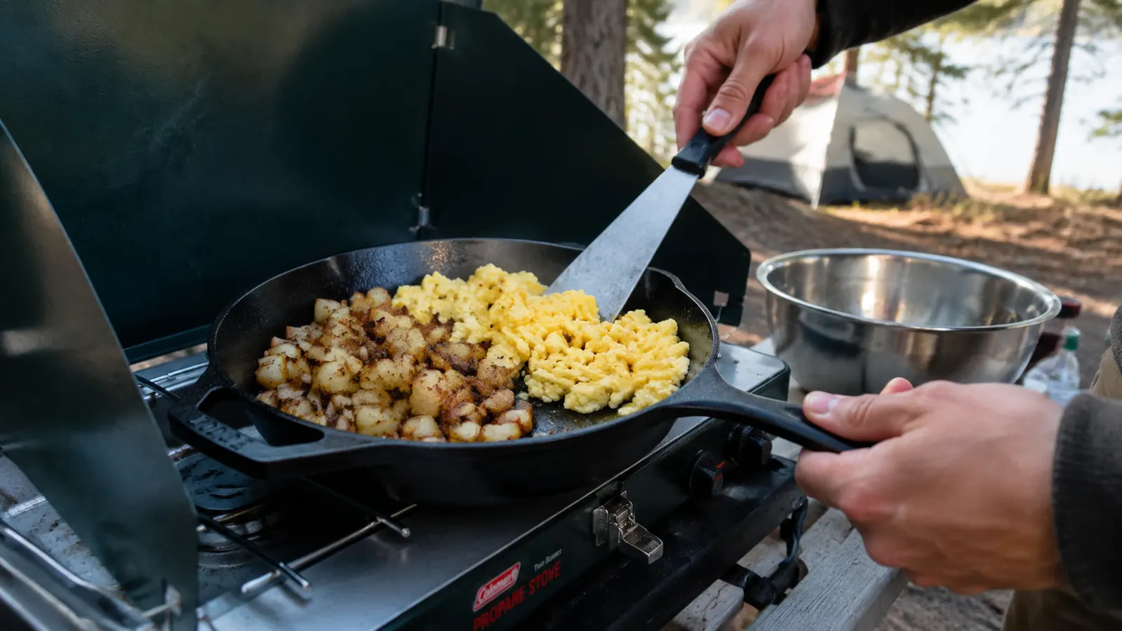 Hands cook with a Lodge cast iron skillet on a two-burner camp stove, lifting browned potatoes and scrambled eggs with a metal spatula